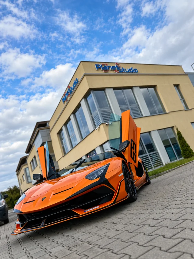 Pomarańczowe Lamborghini z uniesionymi drzwiami przed Planet Studio.
